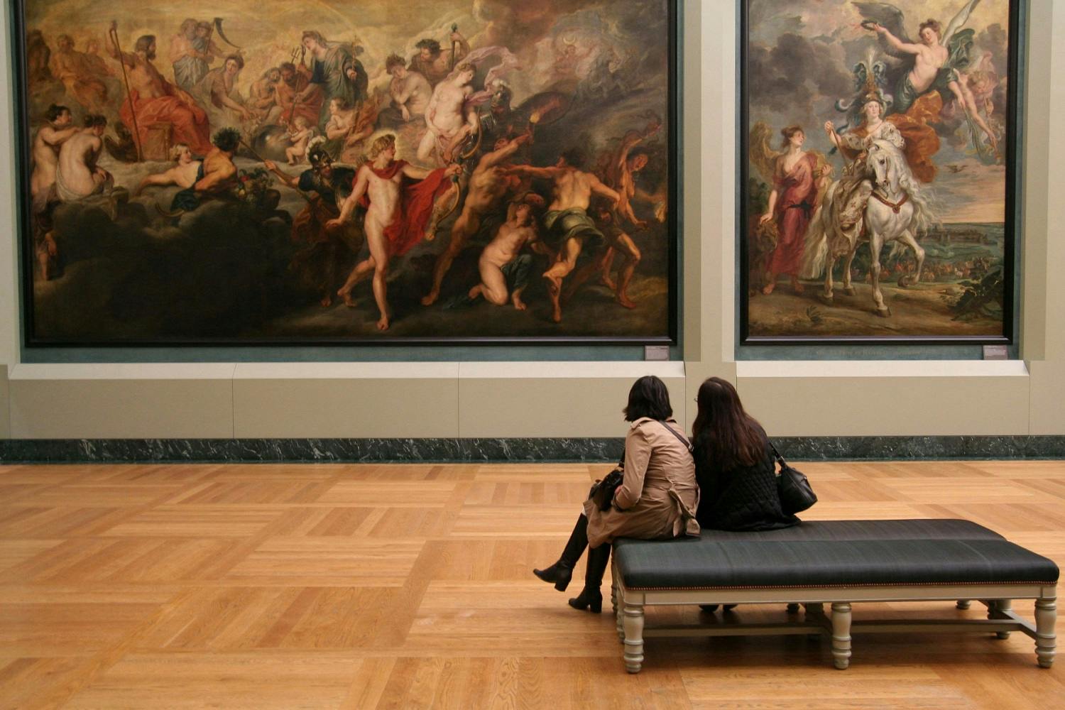 Two women sitting on a bench in an art gallery, contemplating large classical paintings depicting mythological scenes.