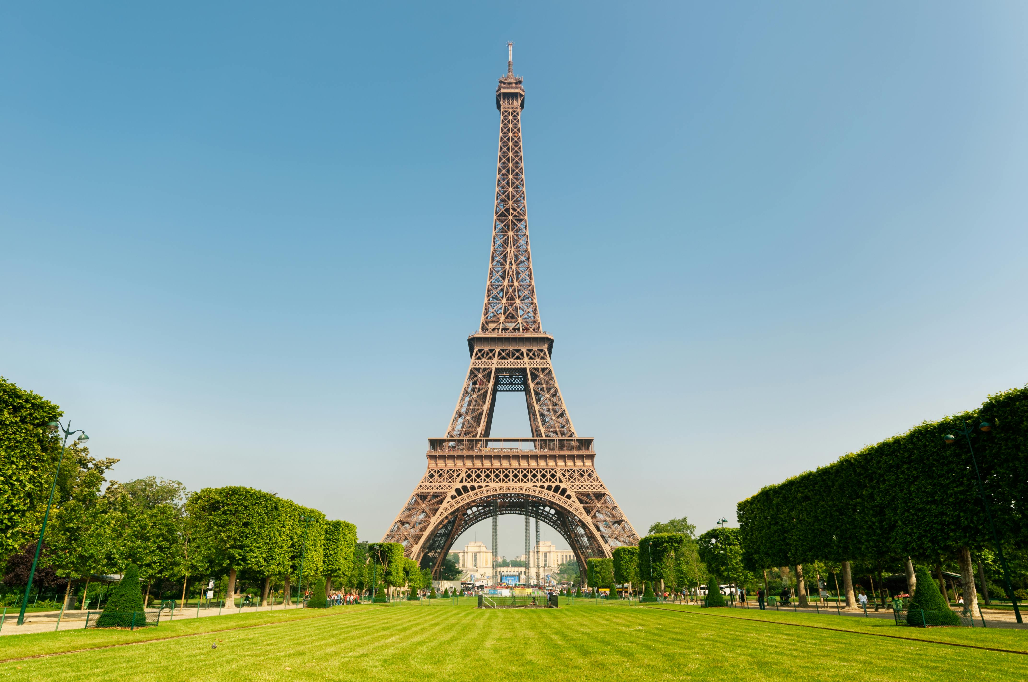 The Eiffel Tower seen from a grassy park with trimmed trees on both sides under a clear blue sky.