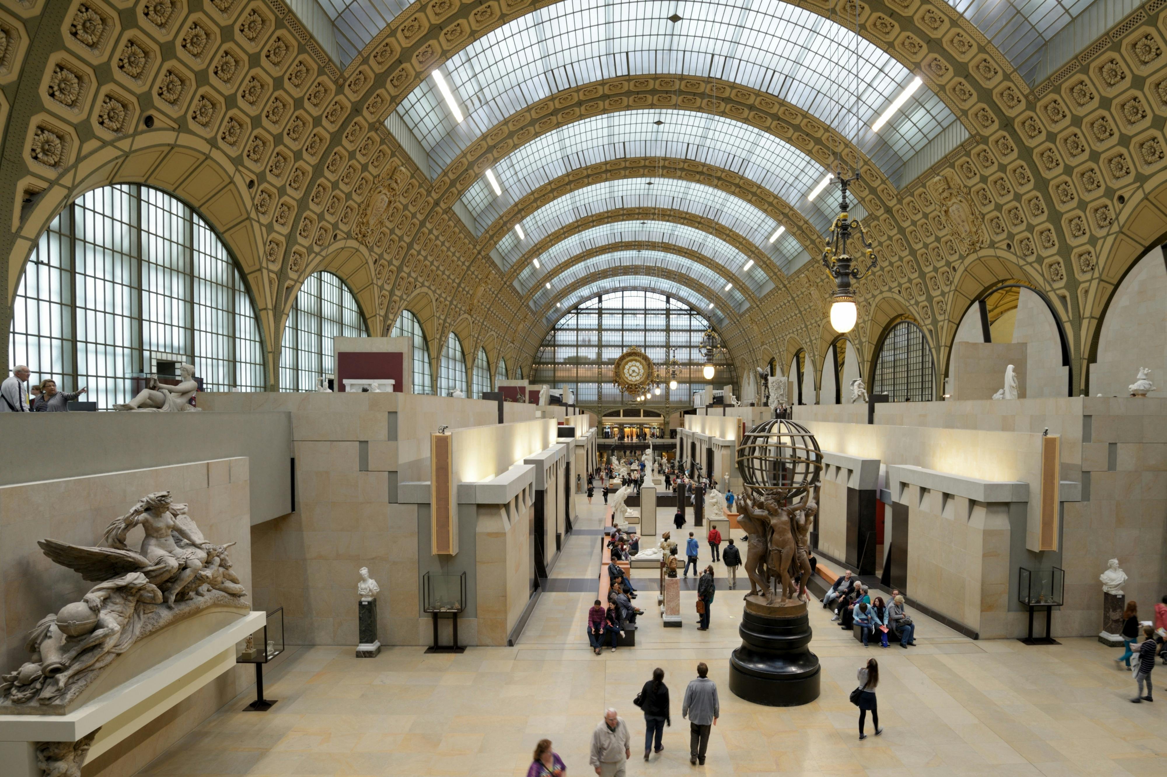 A large museum hall with a vaulted glass ceiling, sculptures, and visitors walking around and sitting on benches.