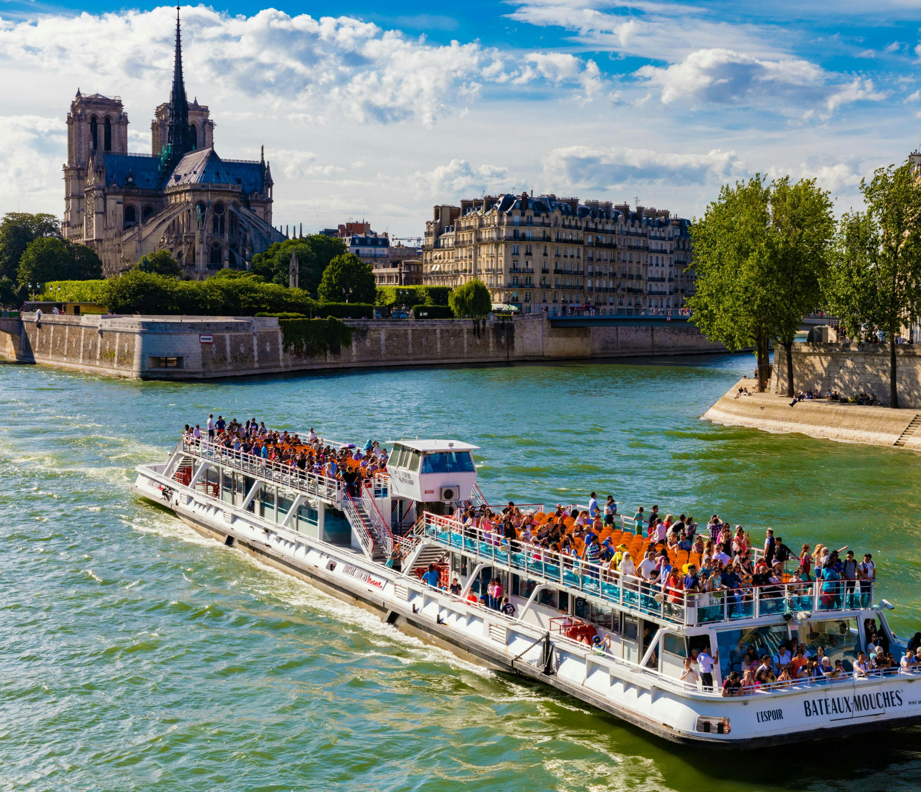 A tourist boat filled with people cruises along a river with Notre-Dame Cathedral and Parisian buildings in the background.