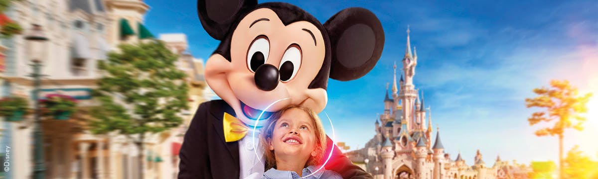 A child smiles at a person dressed as Mickey Mouse, with a fairy-tale castle and colorful buildings in the background.