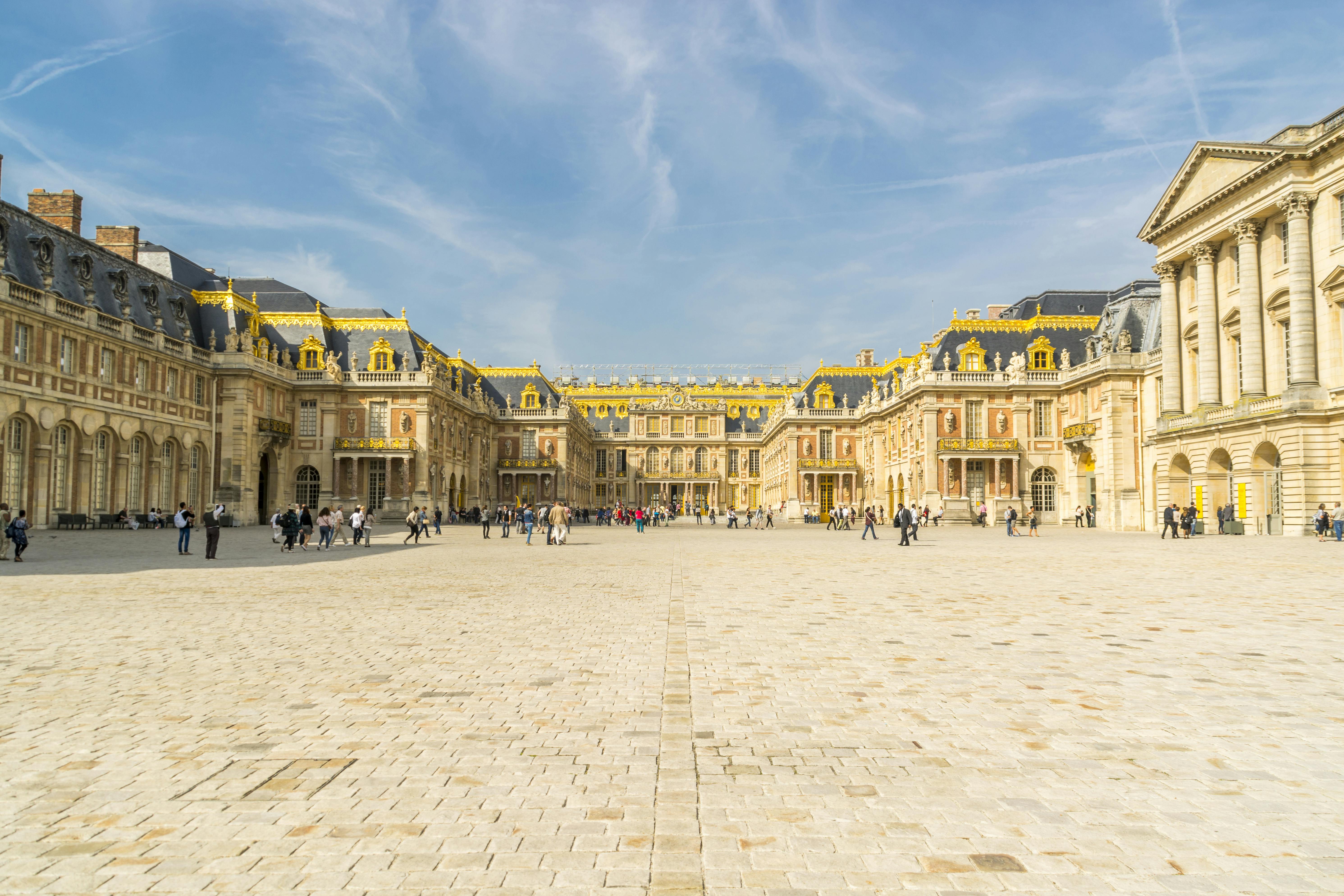 Courtyard of a grand palace with golden decorations on the rooftops. People stroll and take photos on a sunny day.