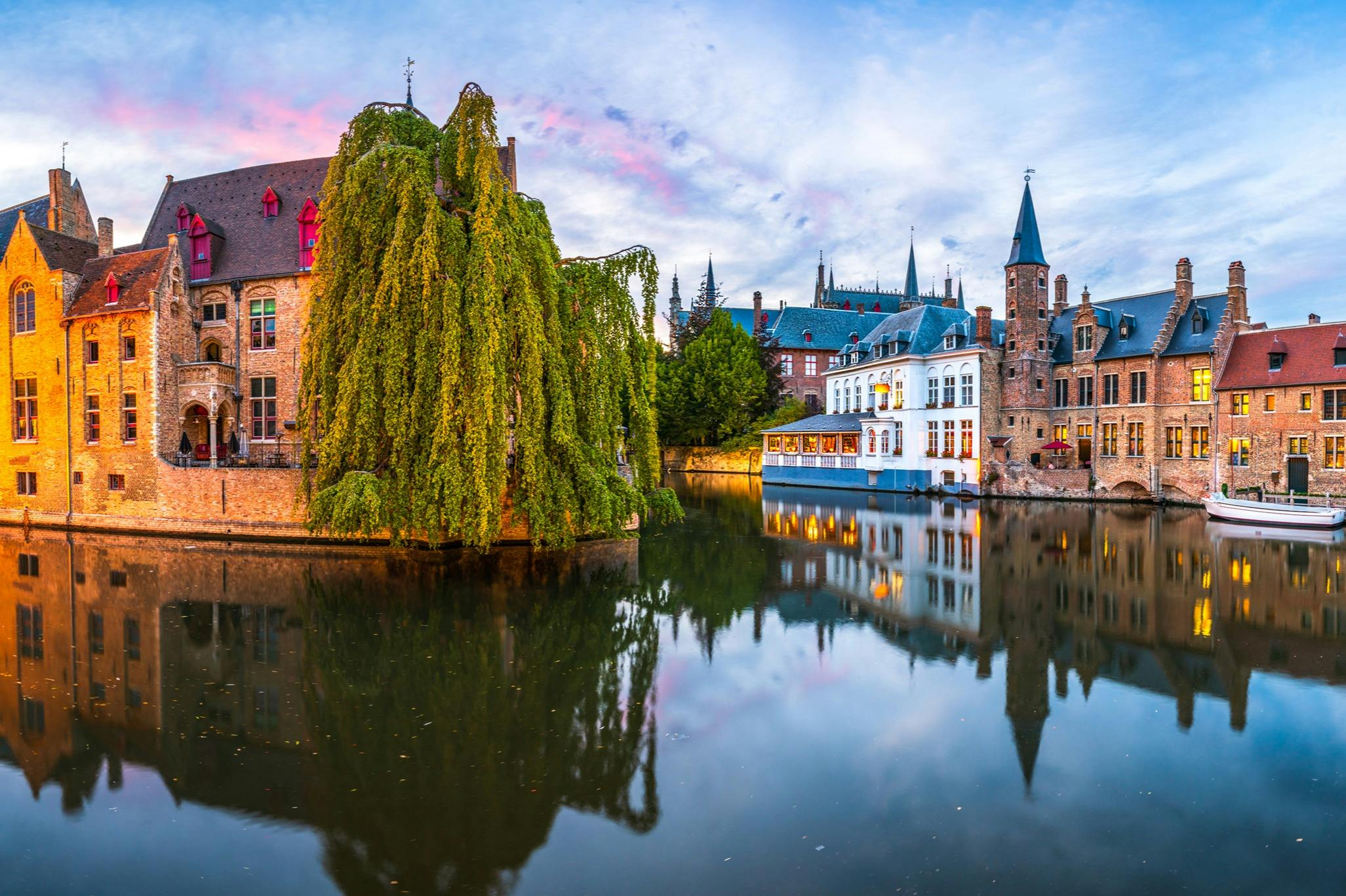 A serene canal scene with historic buildings, a large weeping willow, and spiral rooftops reflected in the water at sunset.