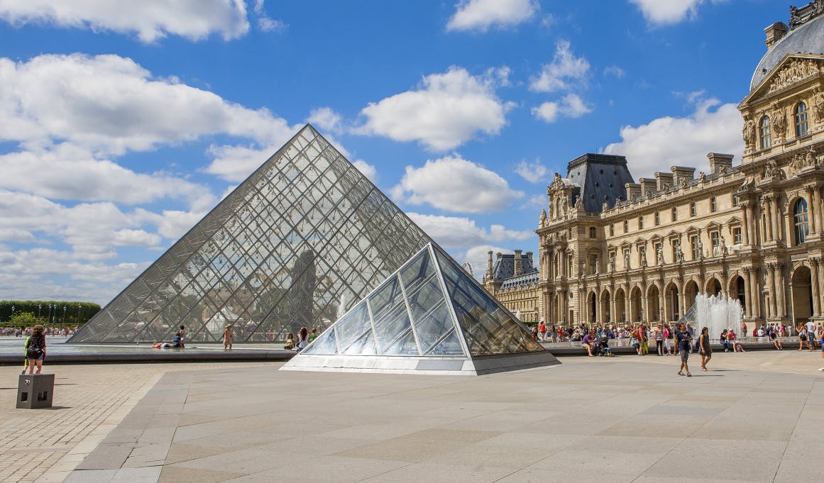 Glass pyramids of the Louvre Museum with people strolling and resting around them, the historic building and a blue sky with clouds in the background.