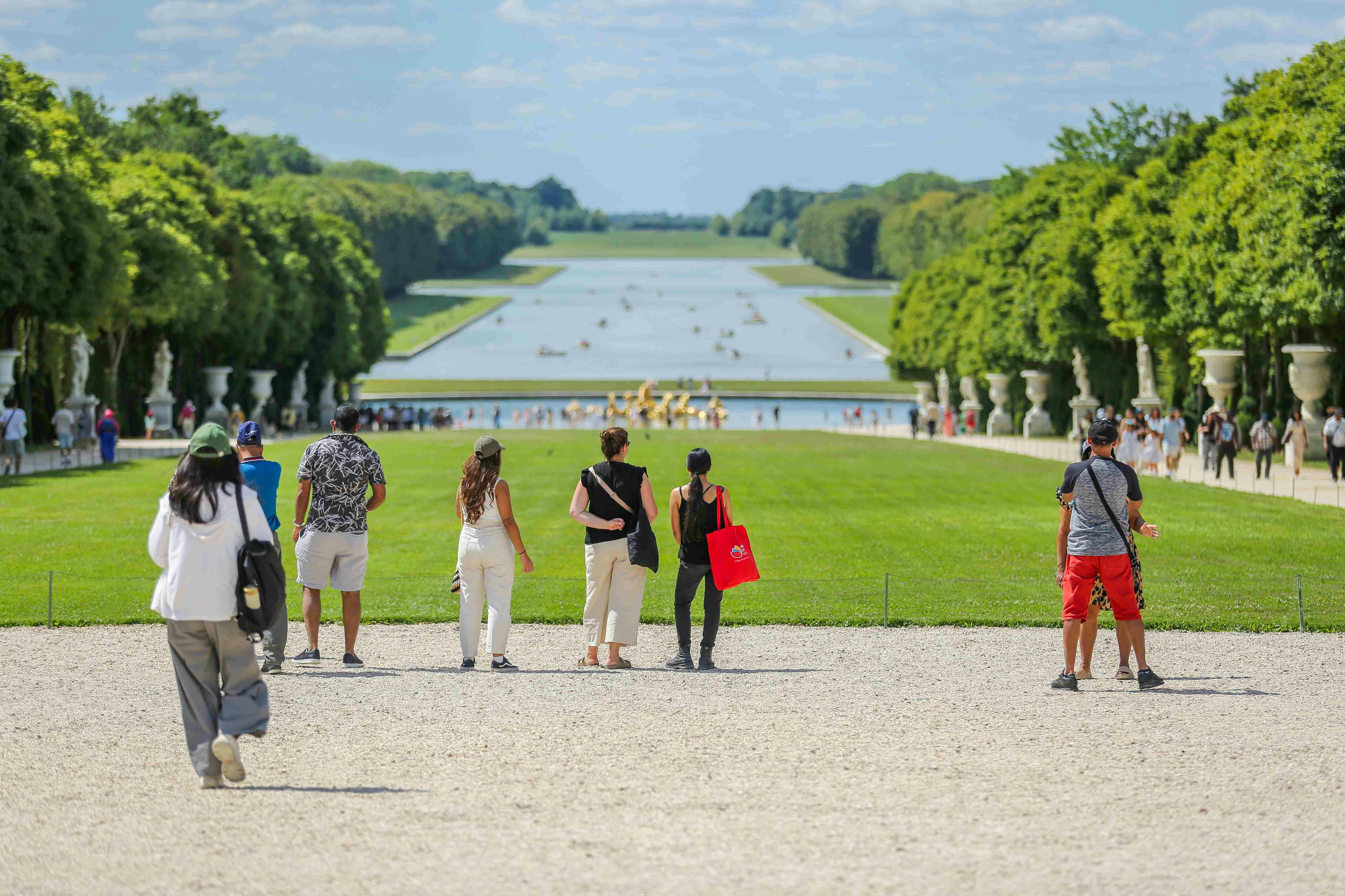 People walking toward a large symmetrical garden with a central canal, lined with trees and distant statues.
